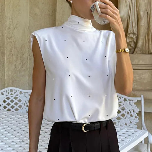 Person wearing a white sleeveless top with black polka dots and black pants, sitting on a white bench.