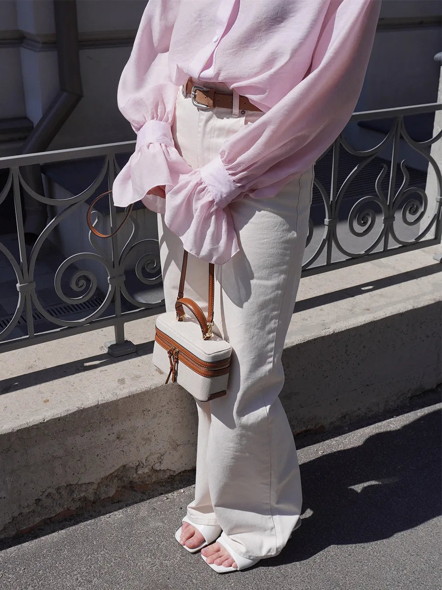 Person wearing a pink blouse and white pants with a small handbag, standing on a street.