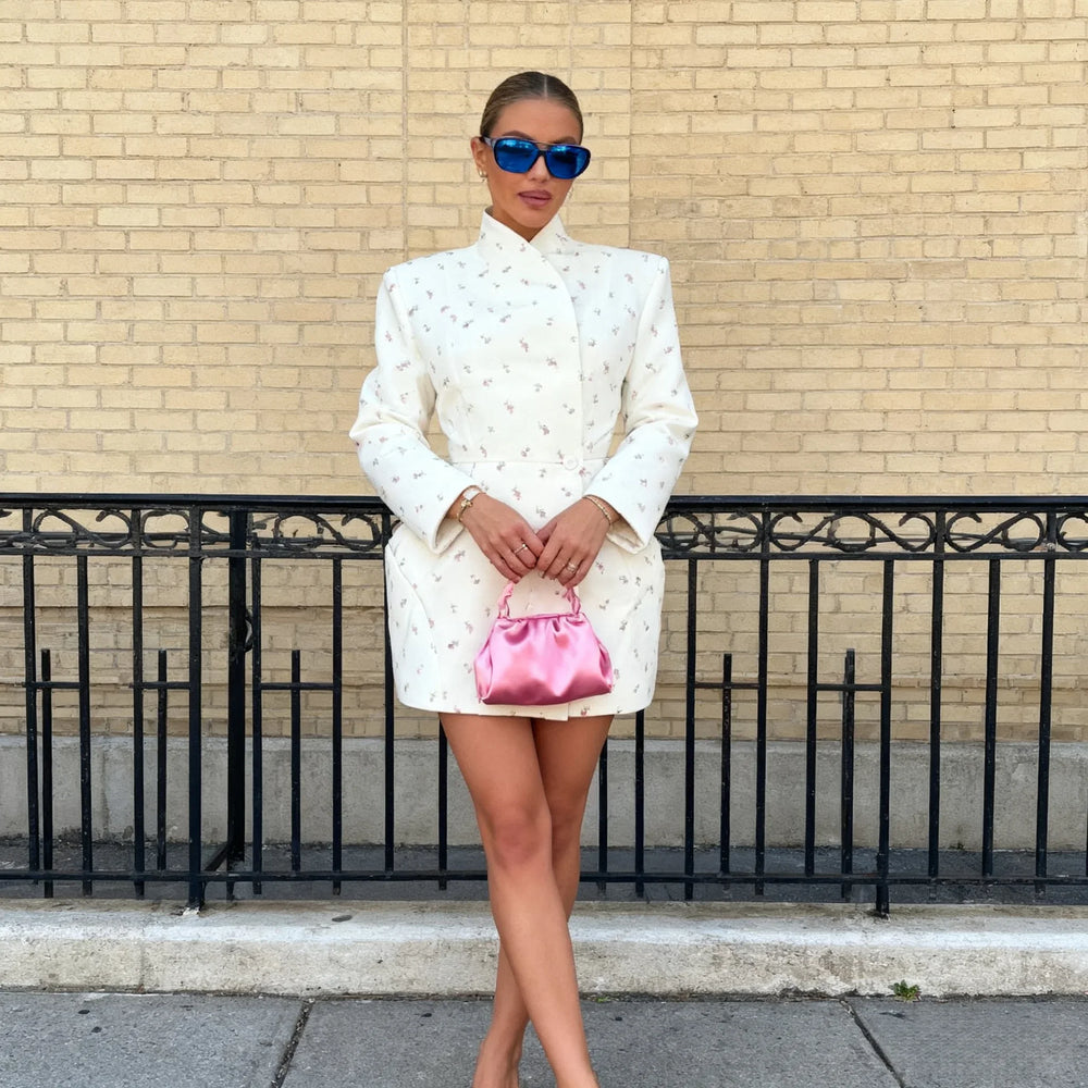Woman in a white outfit holding a pink bag against a brick wall.