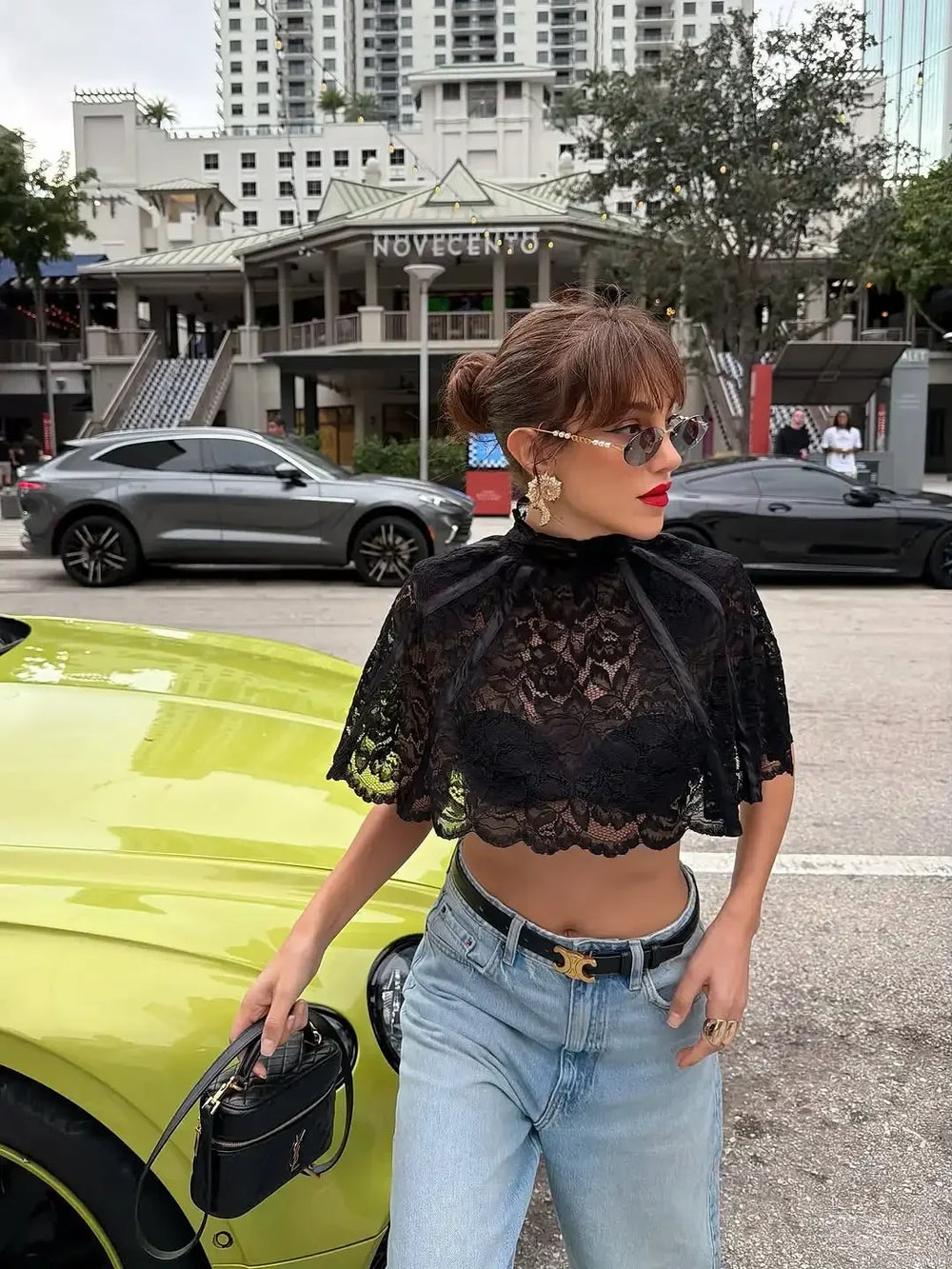 Woman in black lace top and blue jeans standing next to a lime green car with a cityscape background.