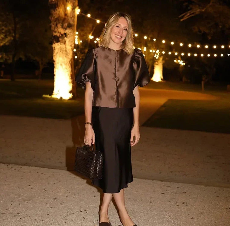 Woman in a brown top and black skirt standing outdoors at night with string lights in the background.