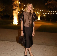 Woman in a brown top and black skirt standing outdoors at night with string lights in the background.