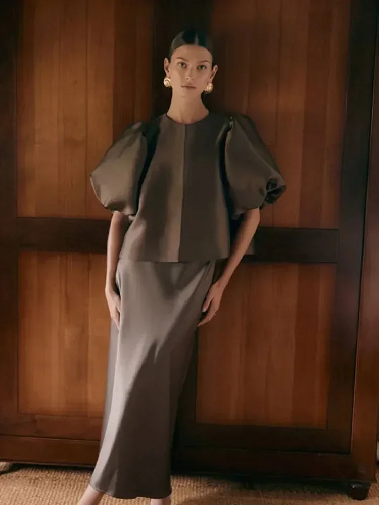 Woman wearing a dark gray outfit standing against a wooden wall.