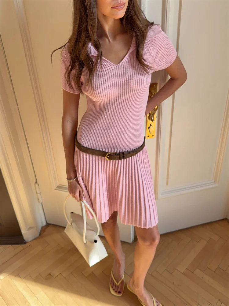 Woman wearing a pink ribbed dress with a belt, standing in a room with wooden flooring.