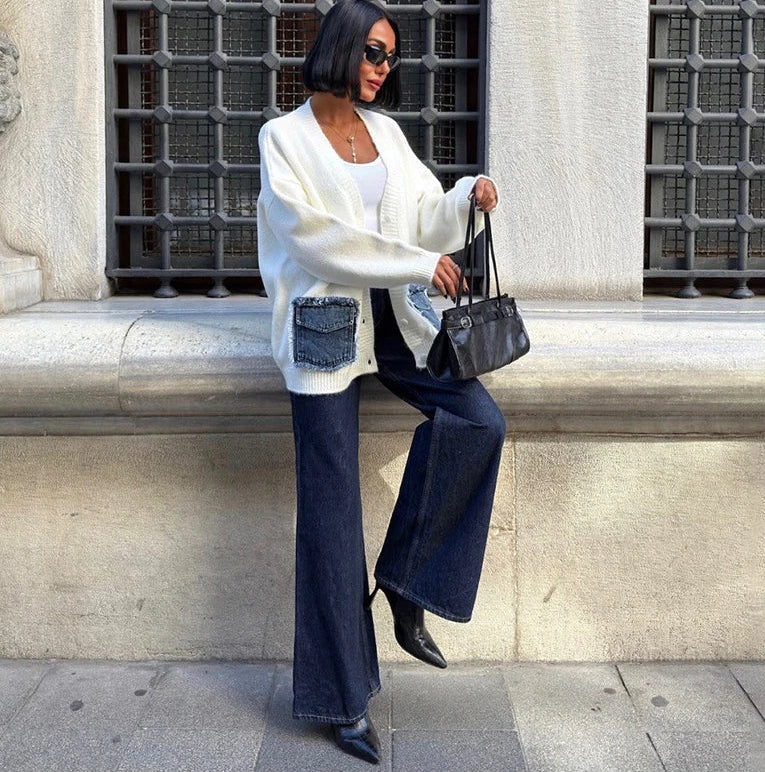 Woman in white cardigan and blue jeans sitting against a stone wall with decorative metalwork.