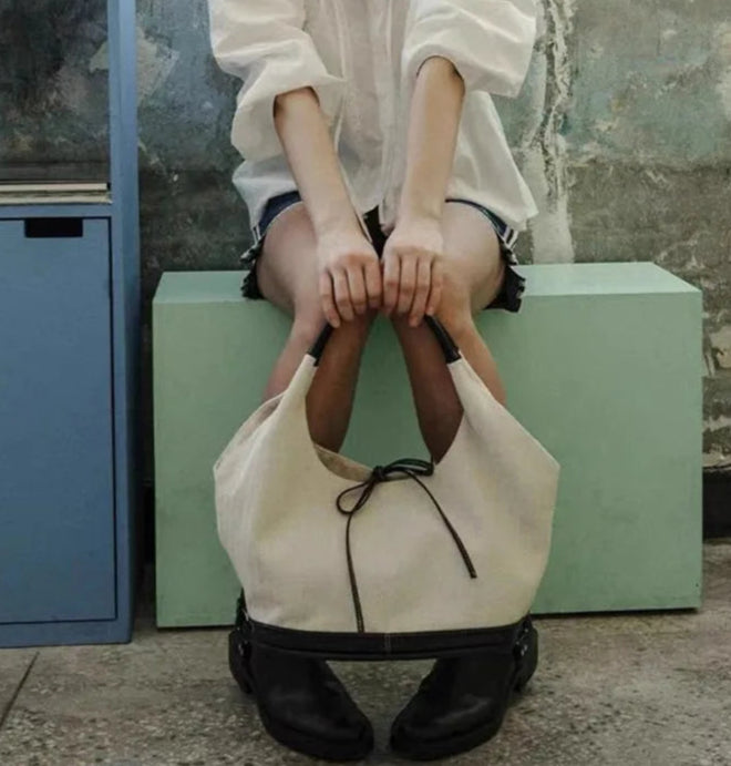 Person sitting on a green box holding a beige handbag with a textured wall and blue cabinet in the background.