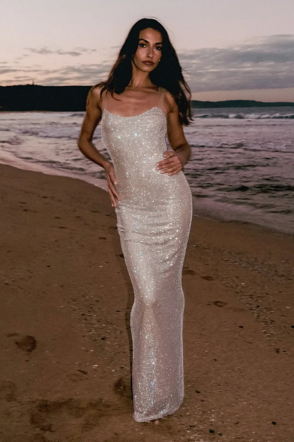 Woman in a sparkly silver dress standing on a beach at sunset.