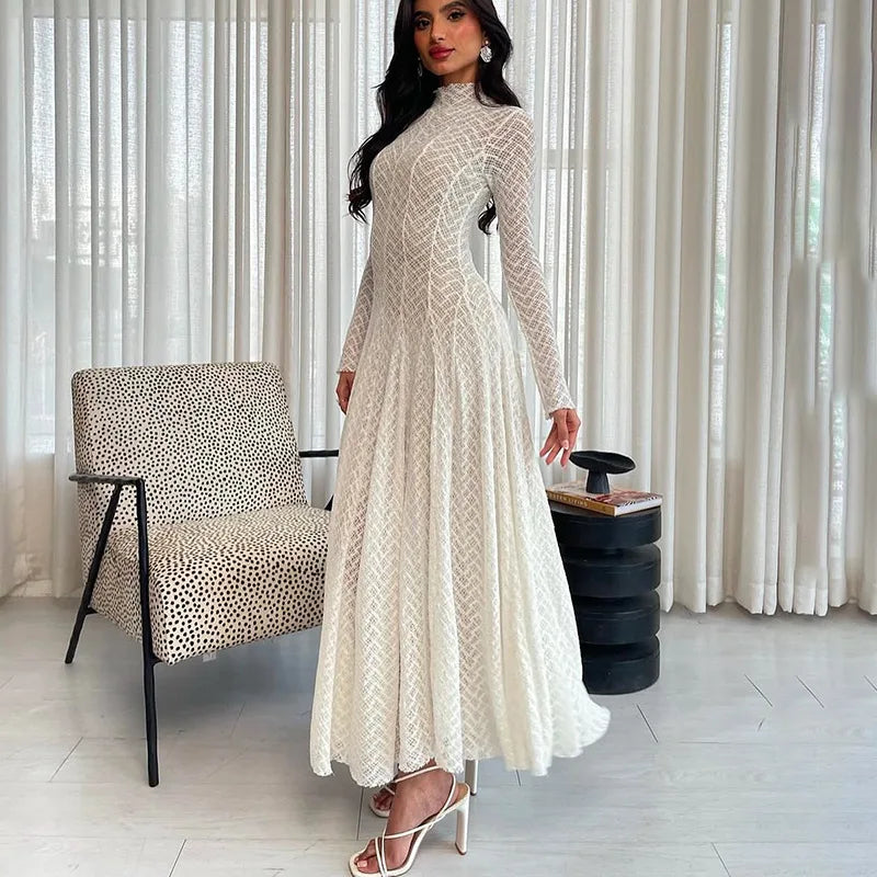 Woman in a long, light-colored lace dress standing in a room with a chair and curtain.