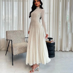 Woman in a long, light-colored lace dress standing in a room with a chair and curtain.