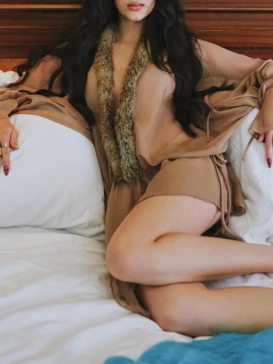 Woman in a beige outfit with a fur stole sitting on a bed.