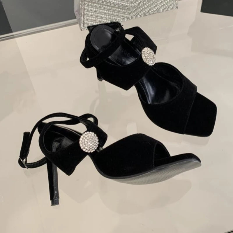 Black stiletto heels with rhinestone embellishment and ankle strap on a reflective surface.