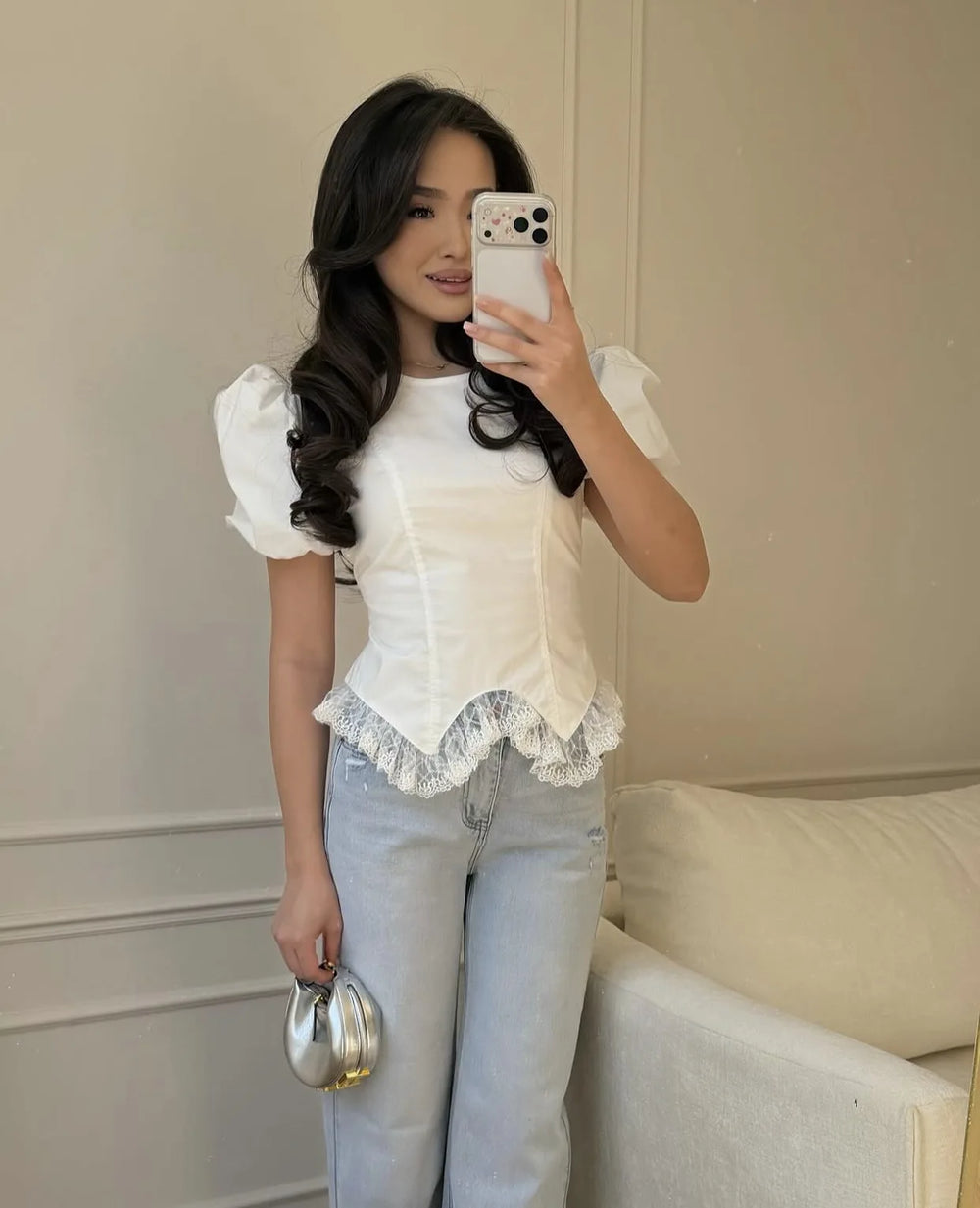 Woman taking a mirror selfie wearing a white blouse with lace details and light blue jeans.