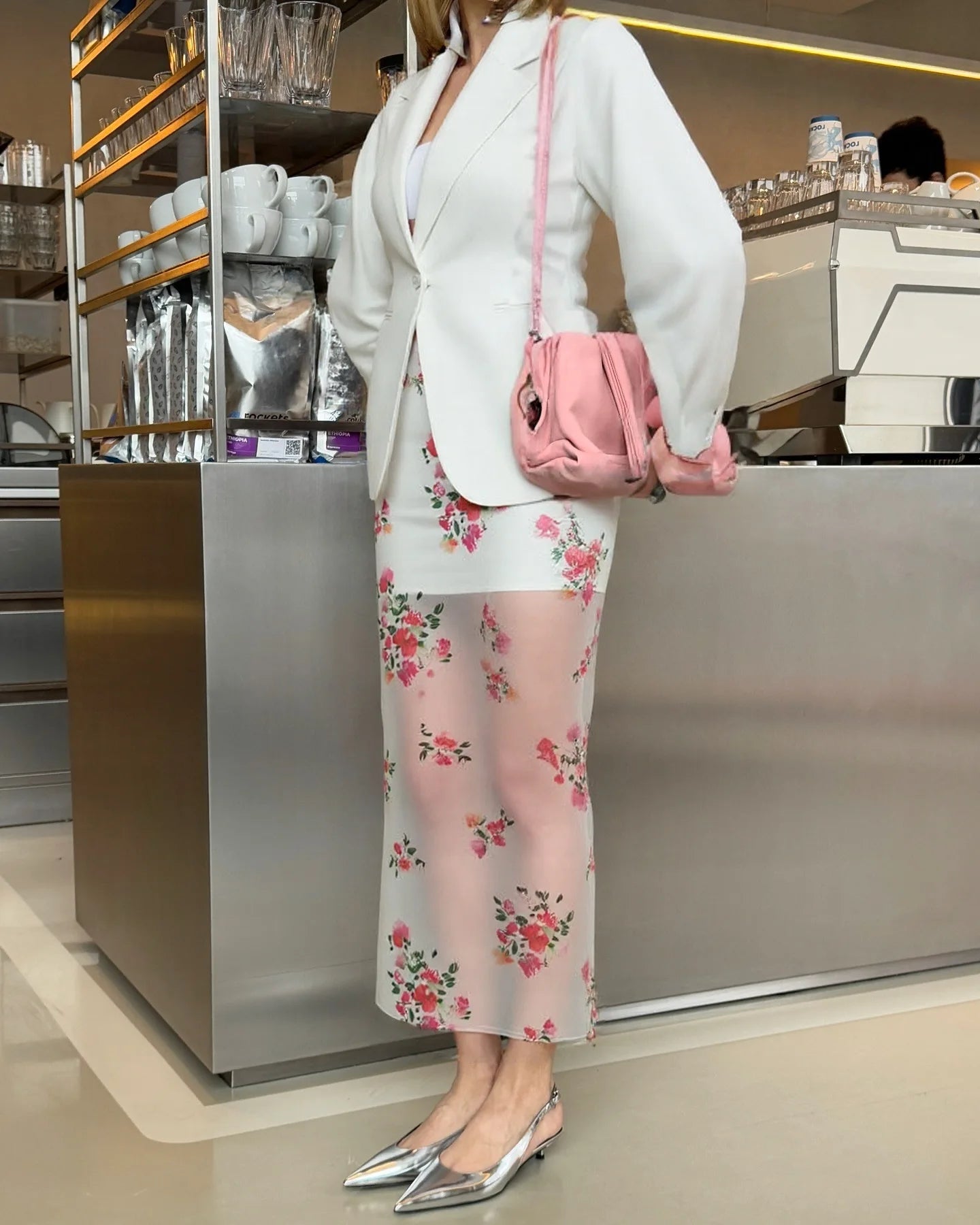 Woman in a white blazer and floral skirt with a pink handbag in a cafe setting