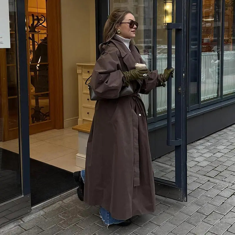 Woman in a long brown coat standing outside a store holding a coffee cup.