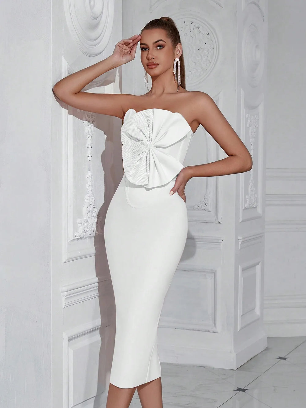 Woman wearing a white strapless dress with a large bow detail against a white decorative wall.