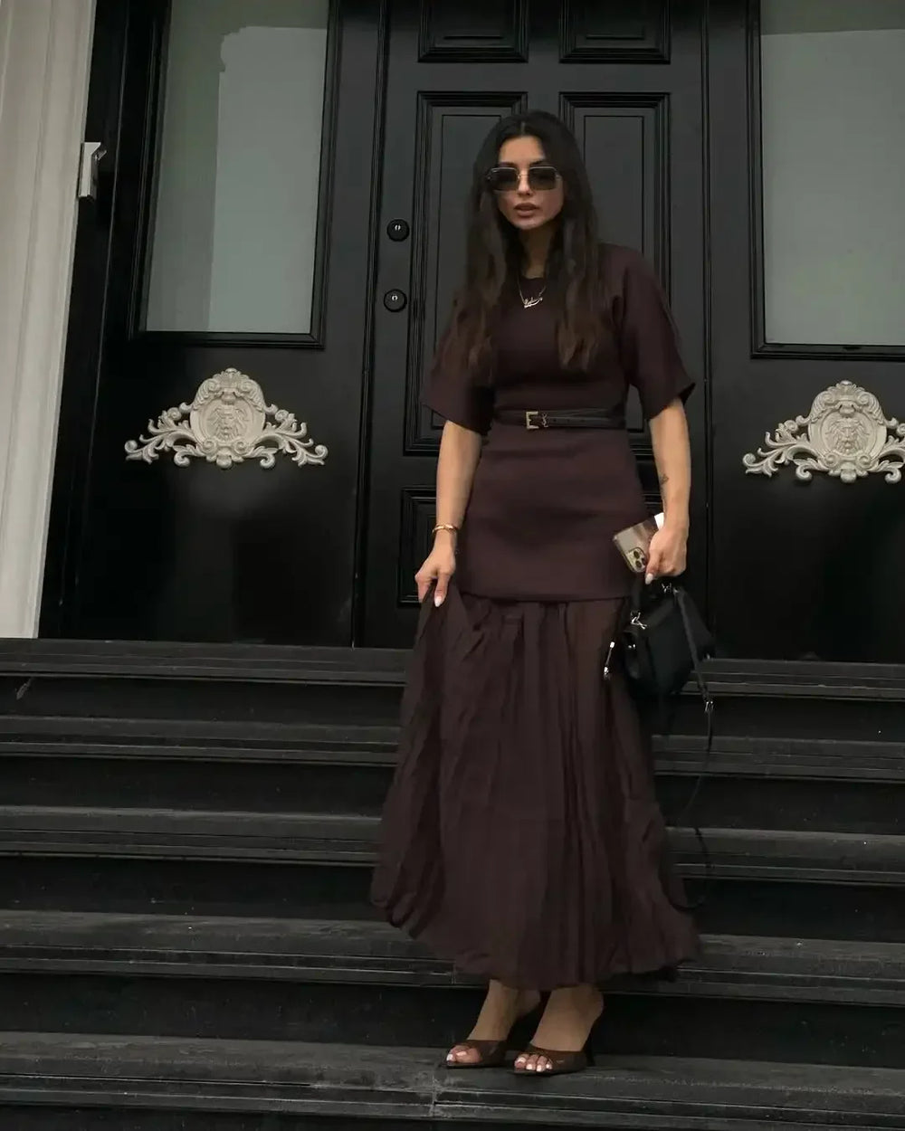 Woman in a dark outfit standing on steps in front of a black door with decorative elements.