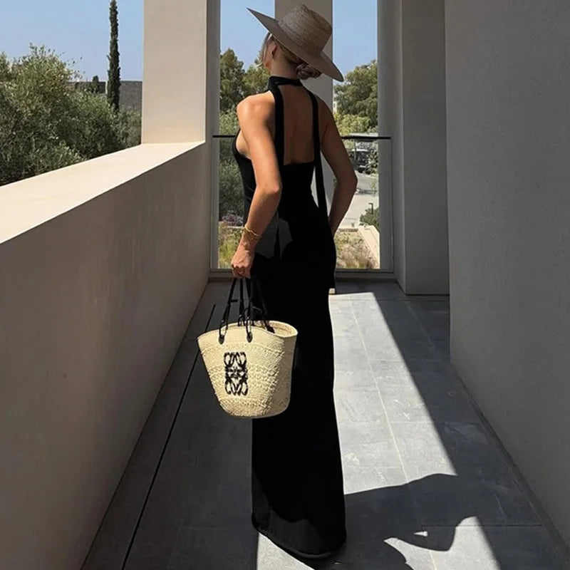 Woman in a black dress and straw hat holding a woven bag on a modern patio.