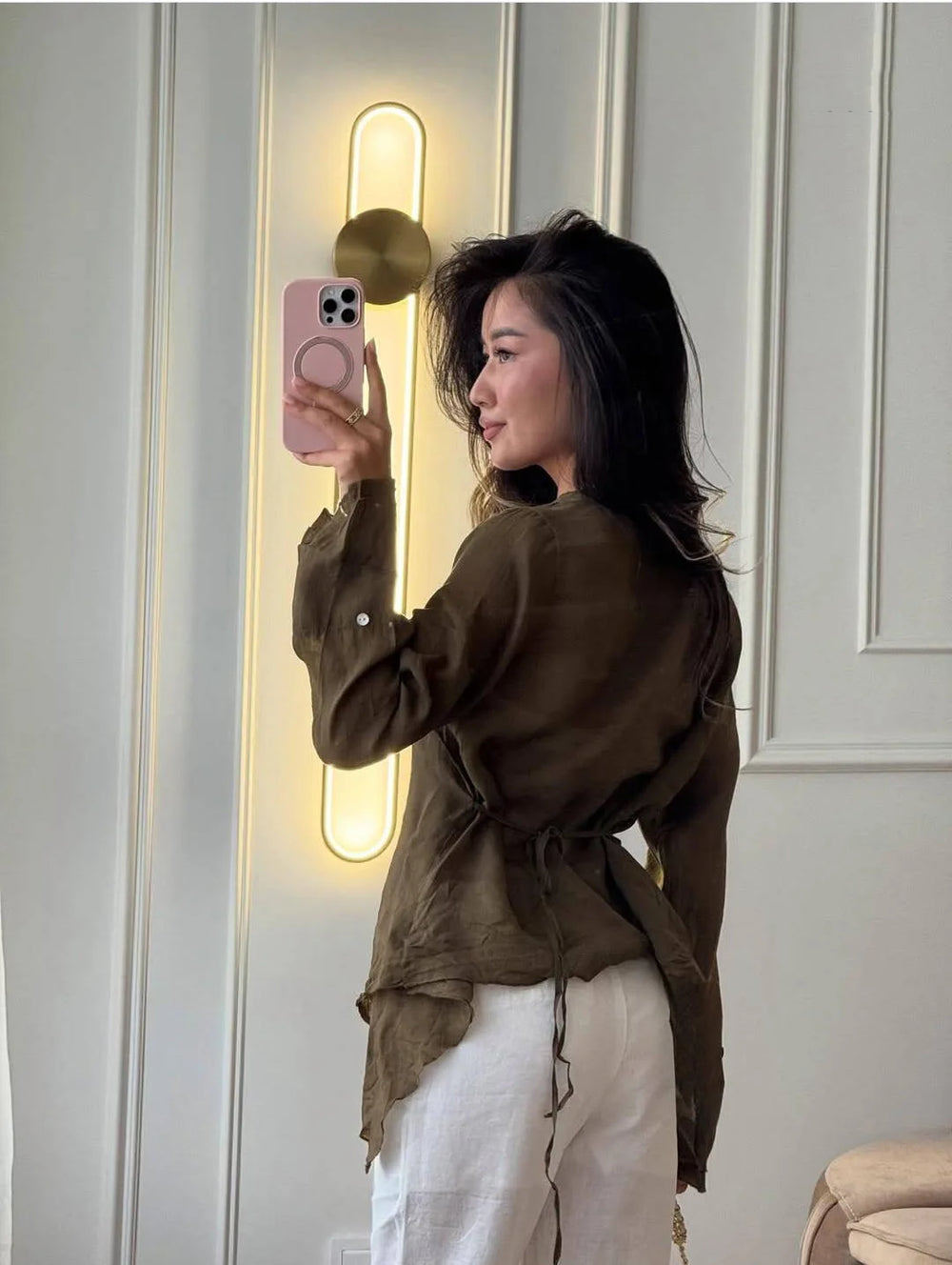 Person taking a mirror selfie wearing a brown top and white pants in a room with a light fixture.