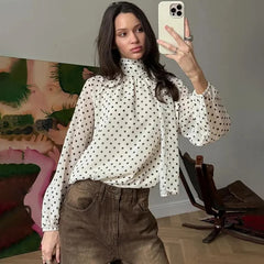Woman taking a mirror selfie wearing a polka dot blouse and brown pants in a room.