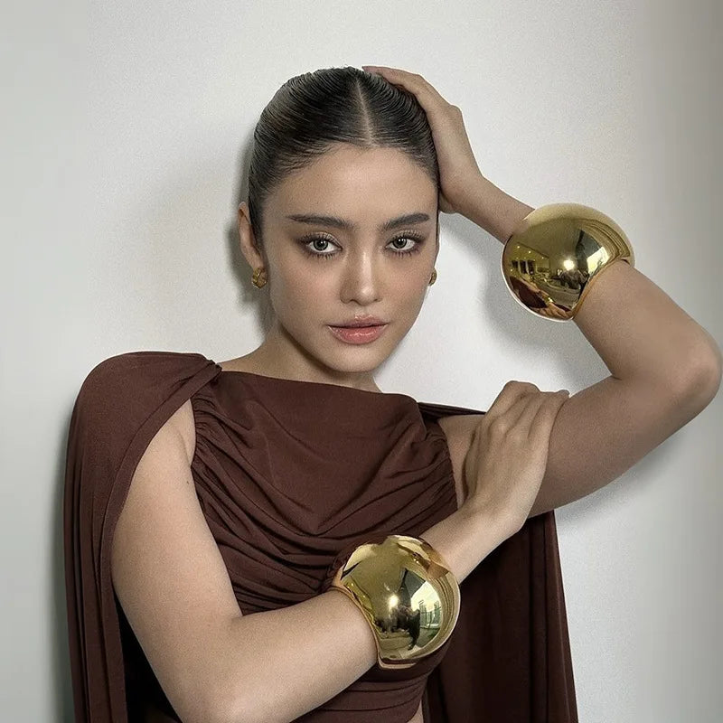 Woman wearing a brown garment and gold arm cuffs against a plain background