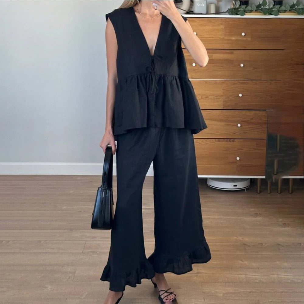 Woman wearing a black sleeveless top and wide-leg pants in a room with wooden flooring and a dresser.