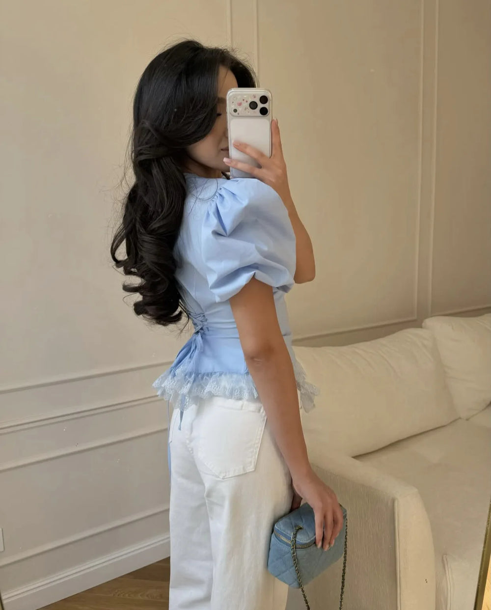 Woman taking a mirror selfie wearing a light blue top and white pants, holding a matching handbag.