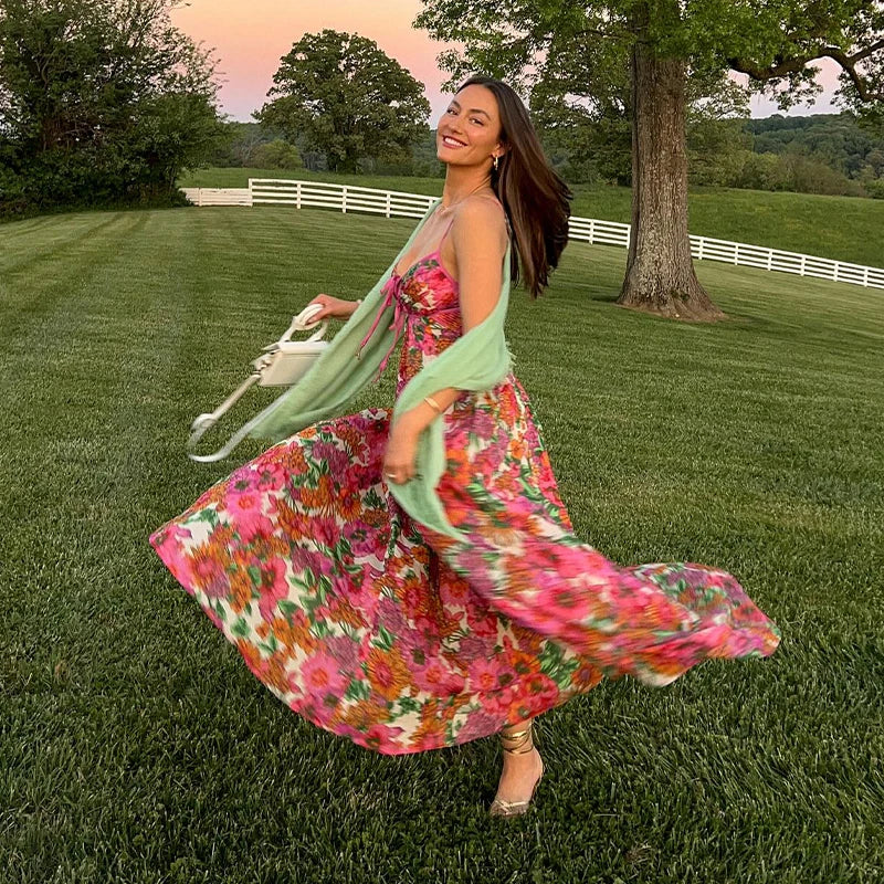Woman in a floral dress standing in a grassy field with trees and a sunset in the background