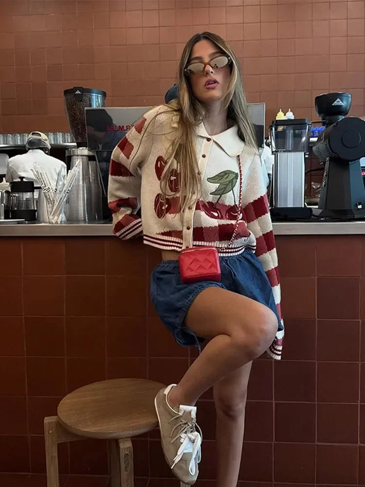 Woman sitting on a stool in a coffee shop wearing a colorful jacket and denim shorts.