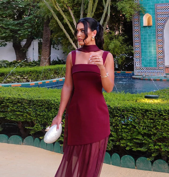 Woman in a burgundy dress standing in a garden with greenery and a decorative wall in the background