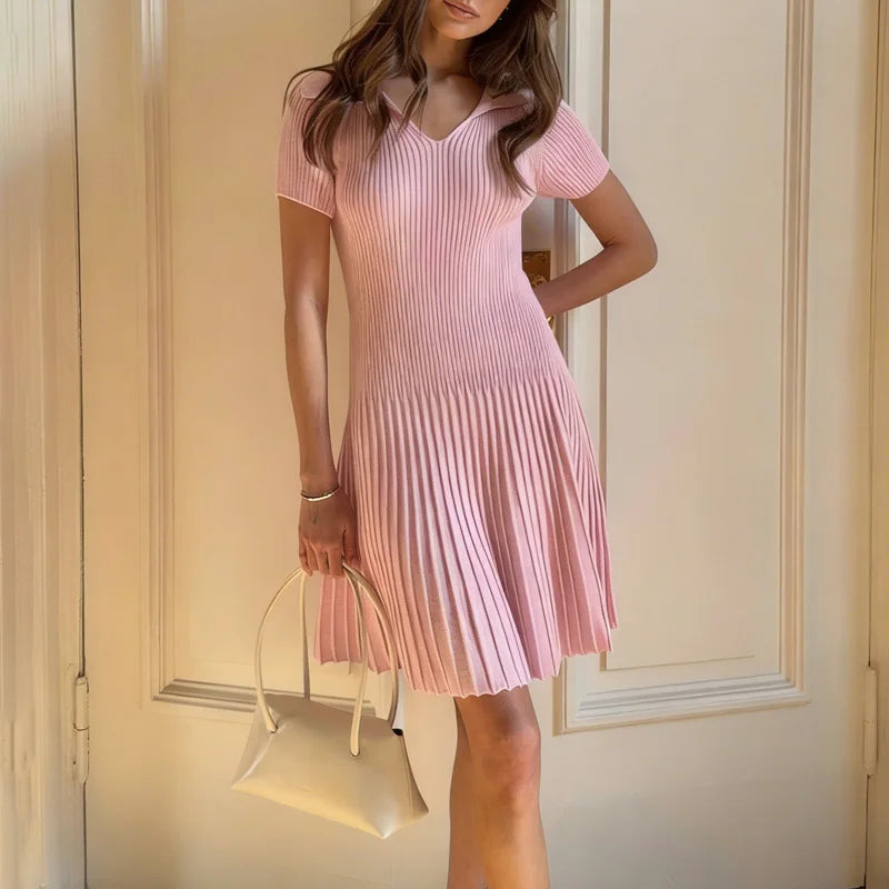 Woman wearing a pink pleated dress holding a beige handbag against a neutral background