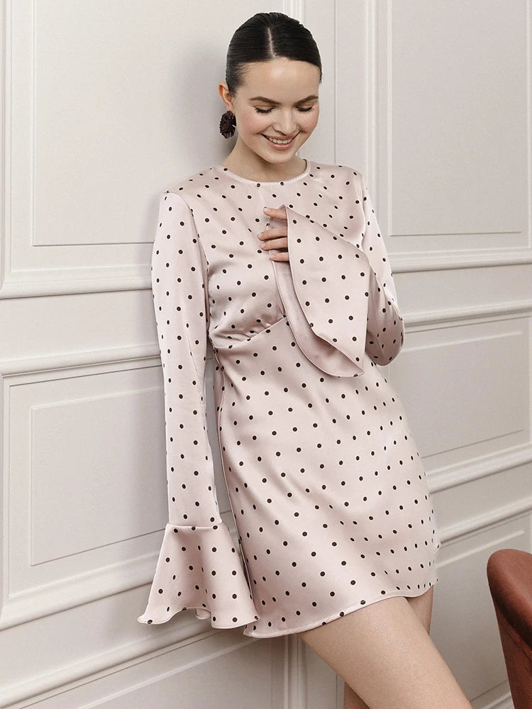 Woman wearing a pink polka dot dress with a ruffled top against a white paneled wall.
