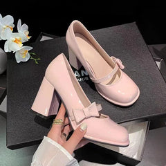 A pair of glossy light pink Mary Jane block-heel pumps, with one held by a hand and the other resting on a black shoebox. White orchids are visible in the top-left corner.