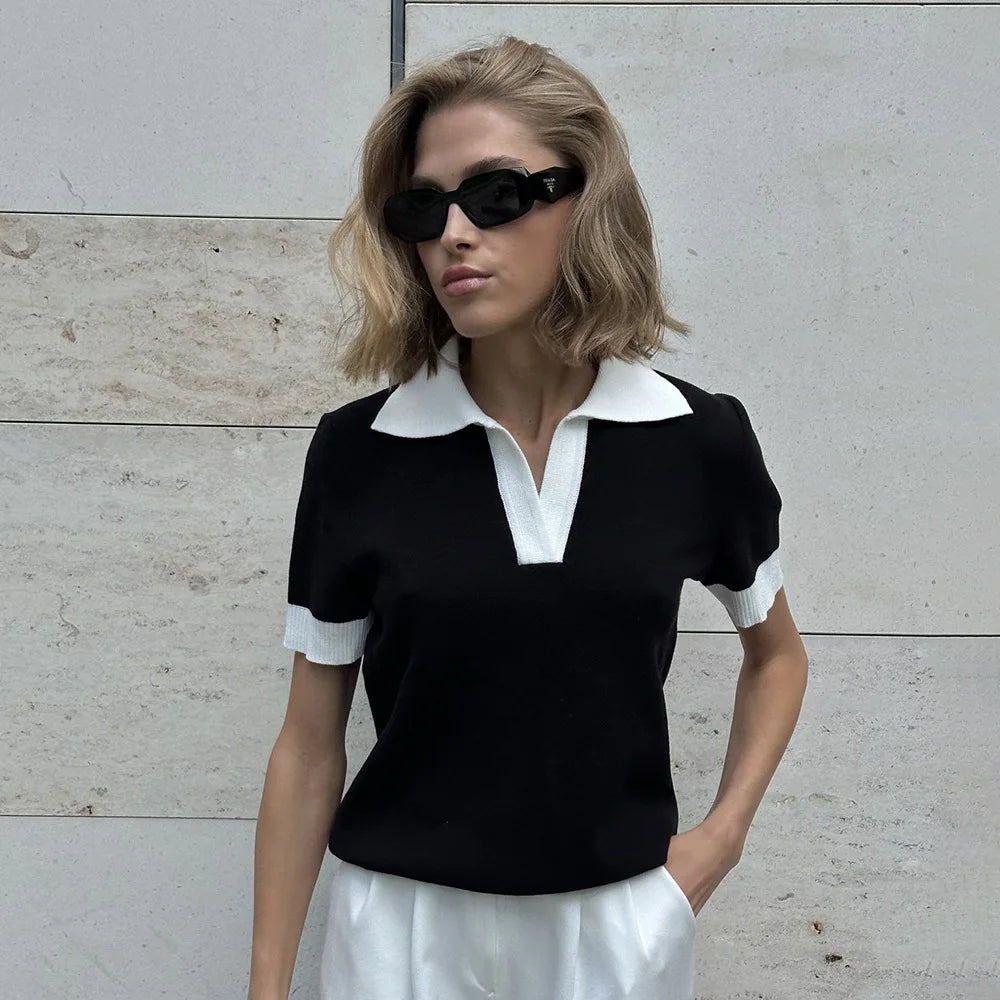 Woman wearing a black and white outfit with sunglasses against a light-colored wall.
