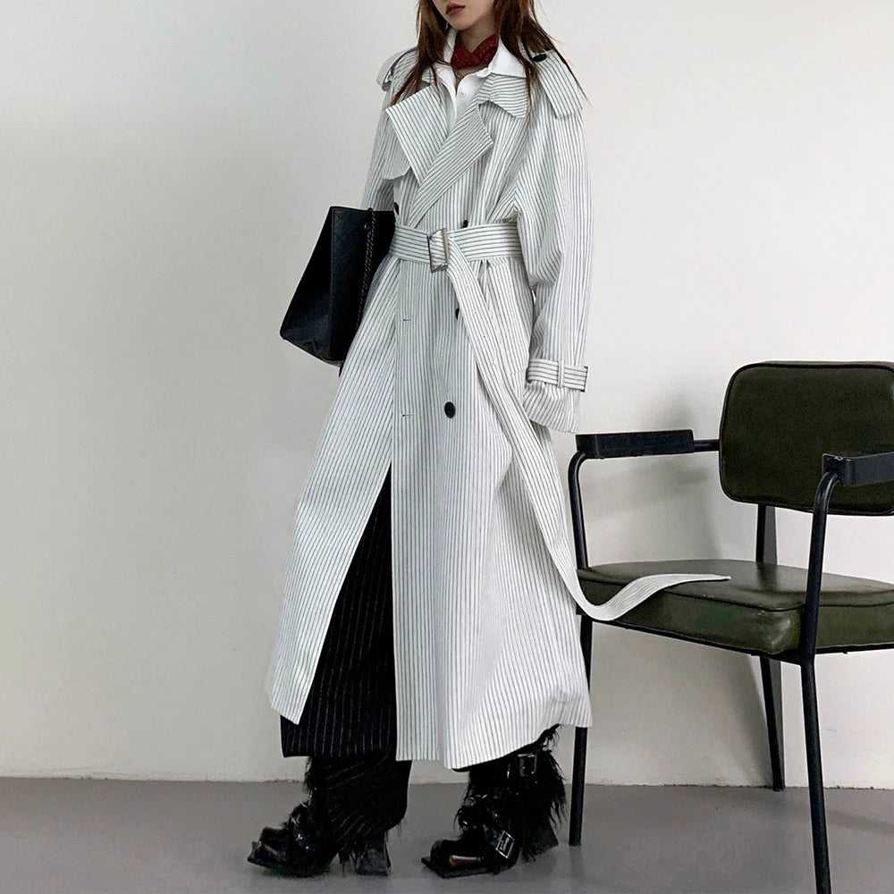 Person wearing a long striped coat standing next to a chair against a plain background