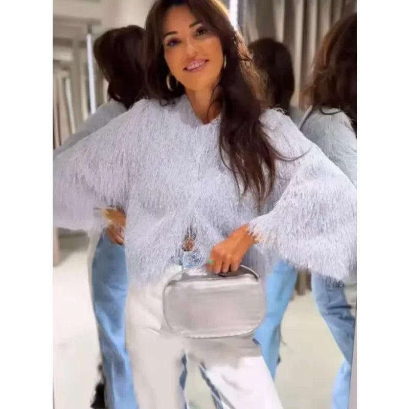Woman in a light blue textured sweater and white pants holding a silver handbag.
