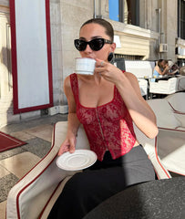 Woman in a red lace top drinking from a white cup outdoors.
