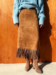 Person wearing a brown suede skirt with fringe and a blue sweater against a wooden background