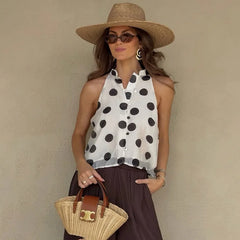 Woman wearing a polka dot sleeveless top and wide-brimmed hat, holding a straw bag against a beige wall.