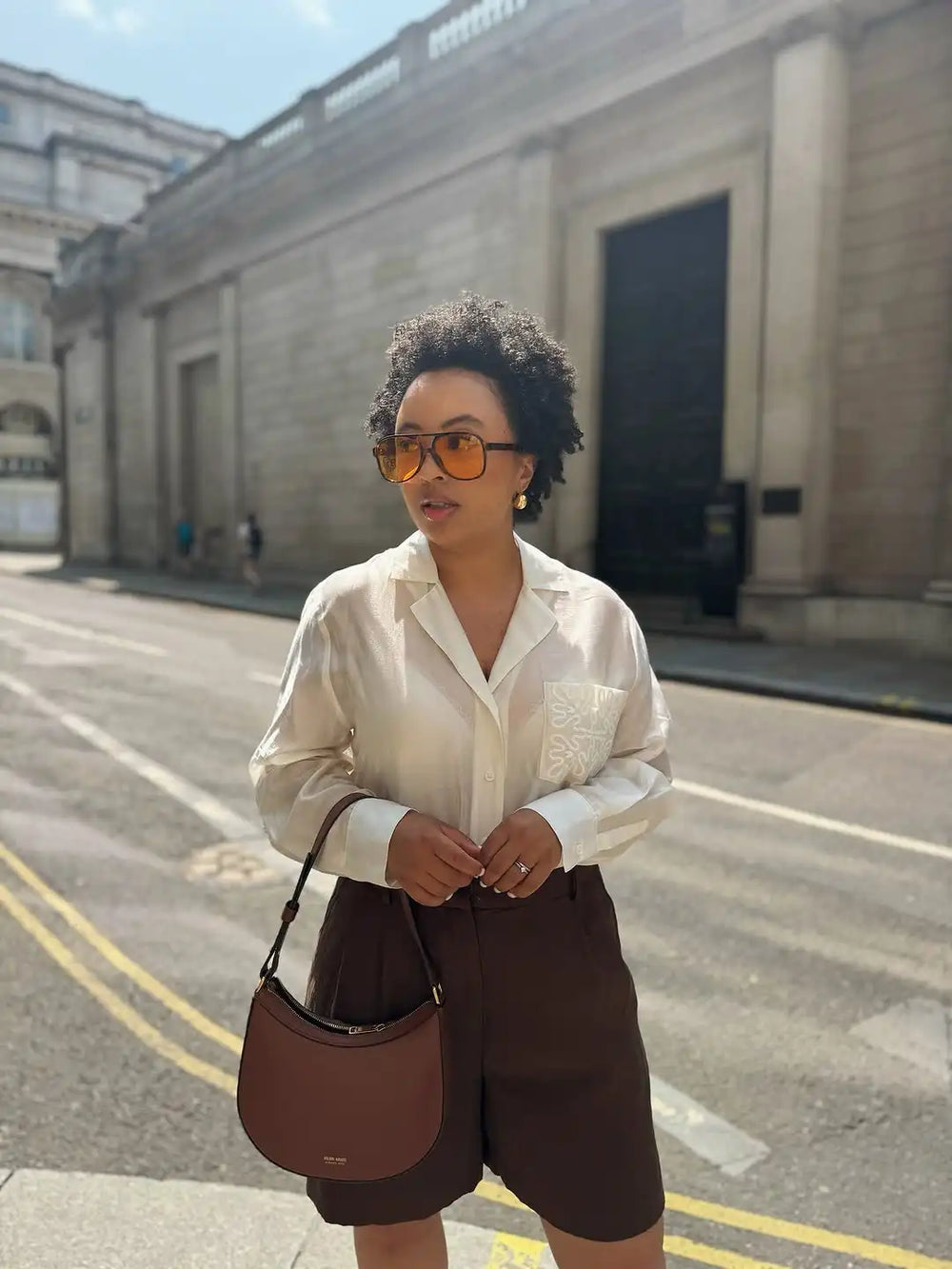 Woman in a cream blouse and brown shorts standing on a street with a building in the background
