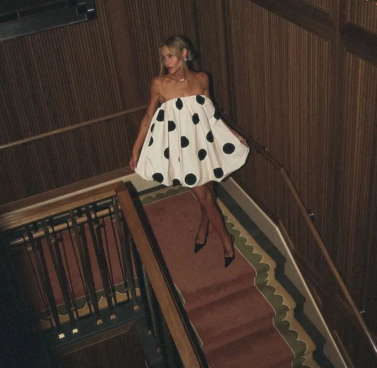 Woman in a polka dot dress walking down a staircase in a wooden-paneled room.