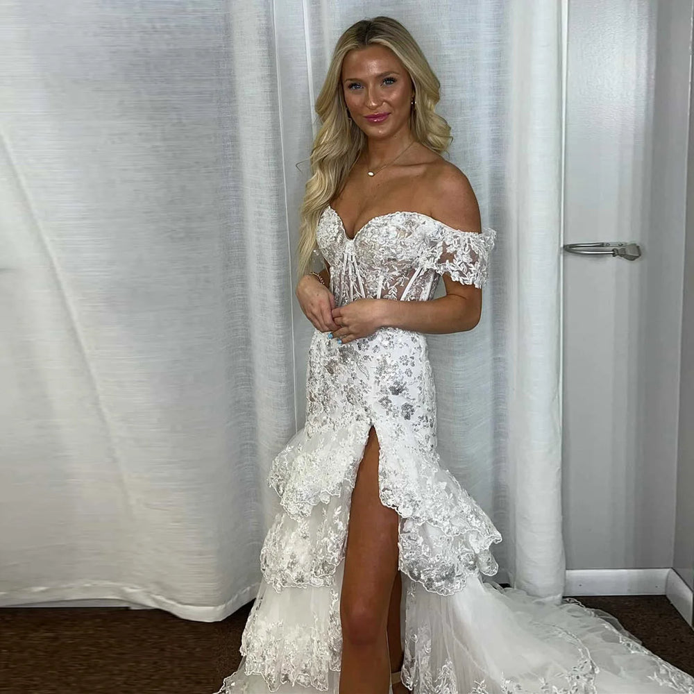 Woman wearing a white lace off-shoulder dress with a high slit, standing against a white curtain.