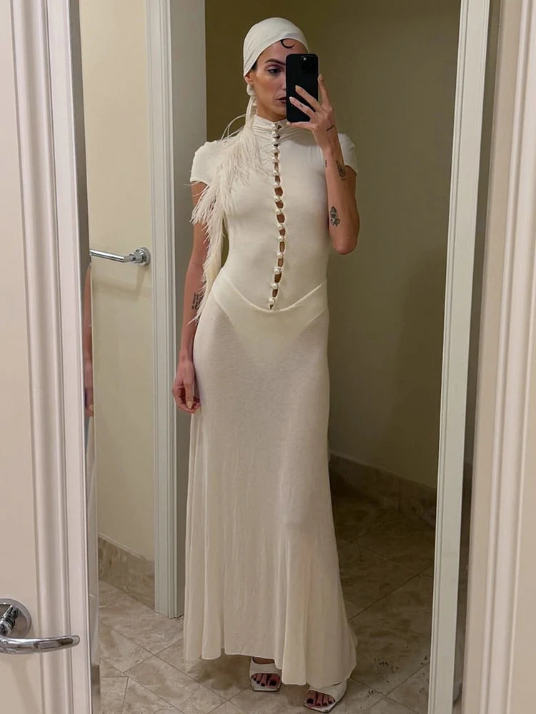 Person wearing a long beige dress with a high neckline and feather details, standing in a bathroom.