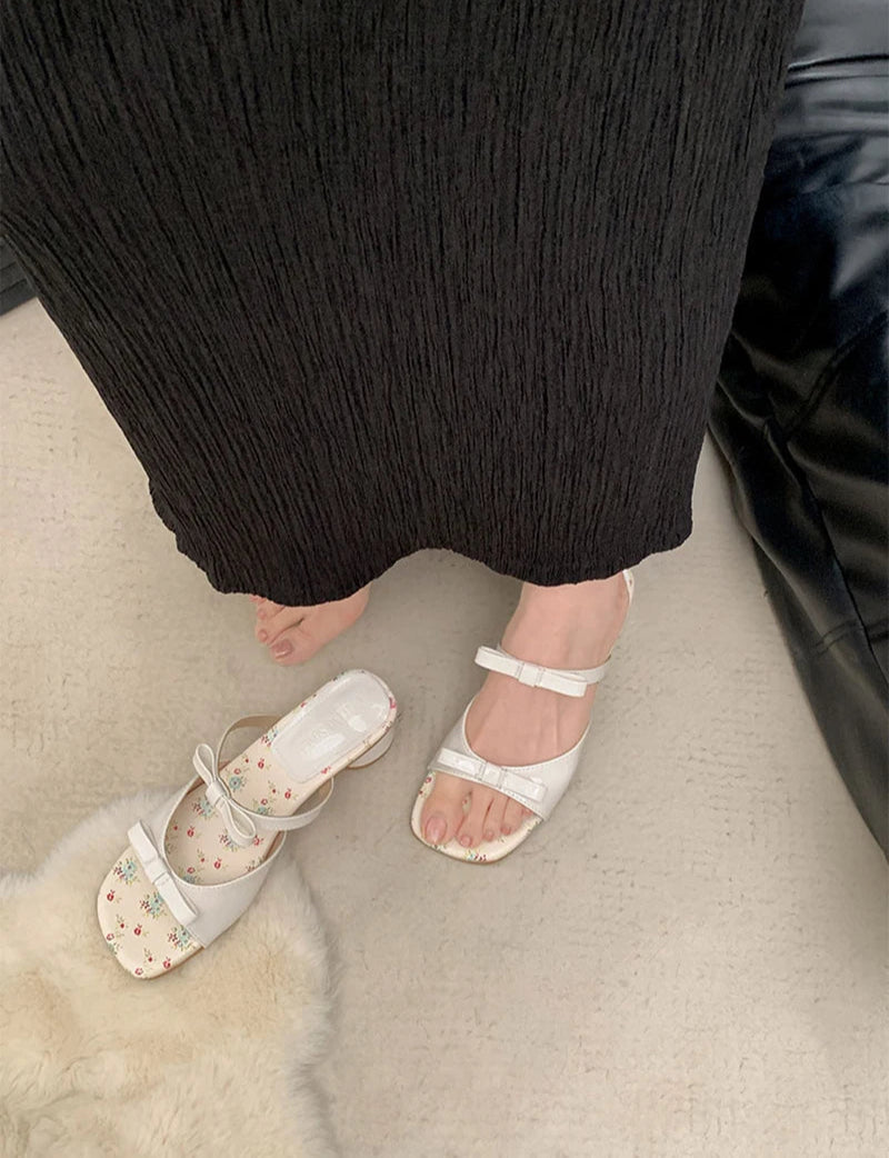 White sandals with floral patterns worn by a person in black pants on a light-colored floor.