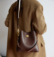 Brown leather handbag with gold accents worn by a person in a brown coat against a neutral background