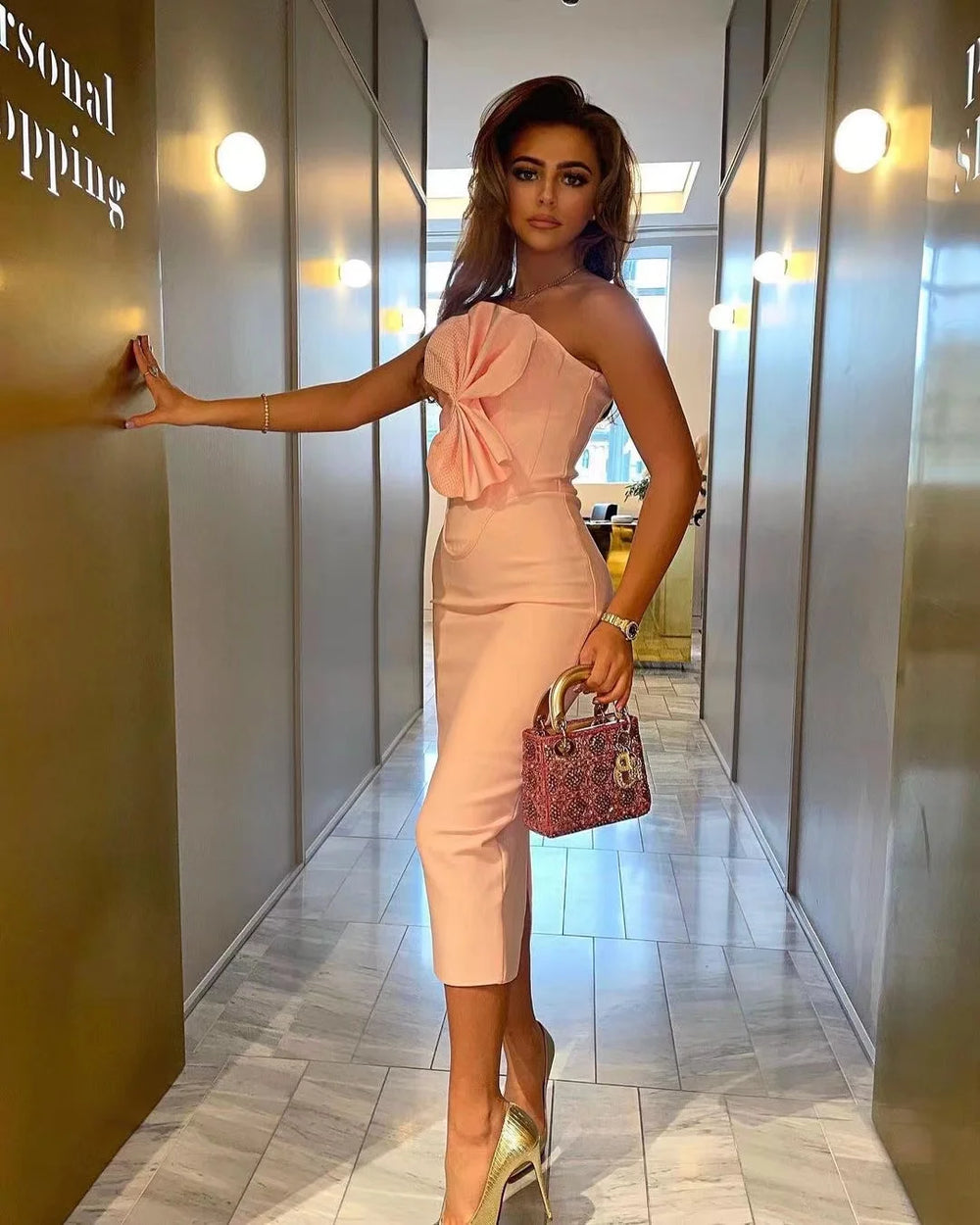 Woman in a pink outfit with a bow detail, holding a small handbag, standing in a modern interior setting.