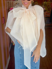 A woman smiles wearing a sheer organza top with a large bow and voluminous sleeves, paired with denim.