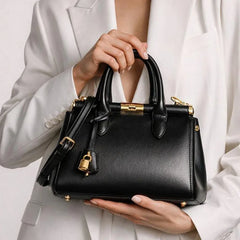 Black structured top-handle satchel bag with gold clasp and lock detail held by a woman.