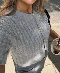 Person wearing a gray cable knit cardigan with gold buttons, holding a drink.