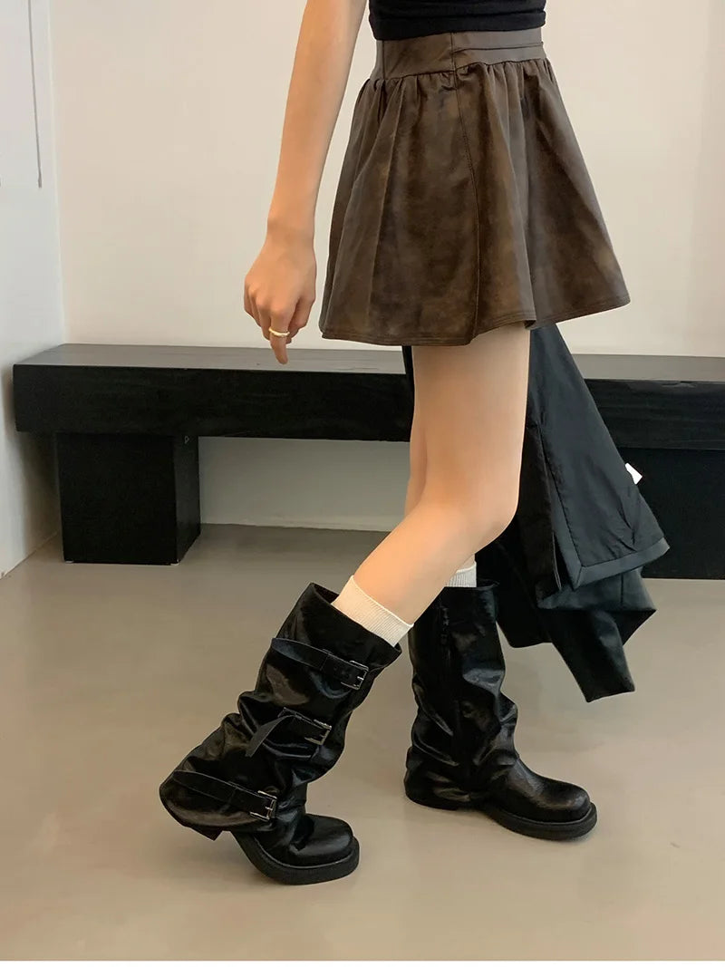 Person wearing a brown skirt and black knee-high boots in a minimal indoor setting.