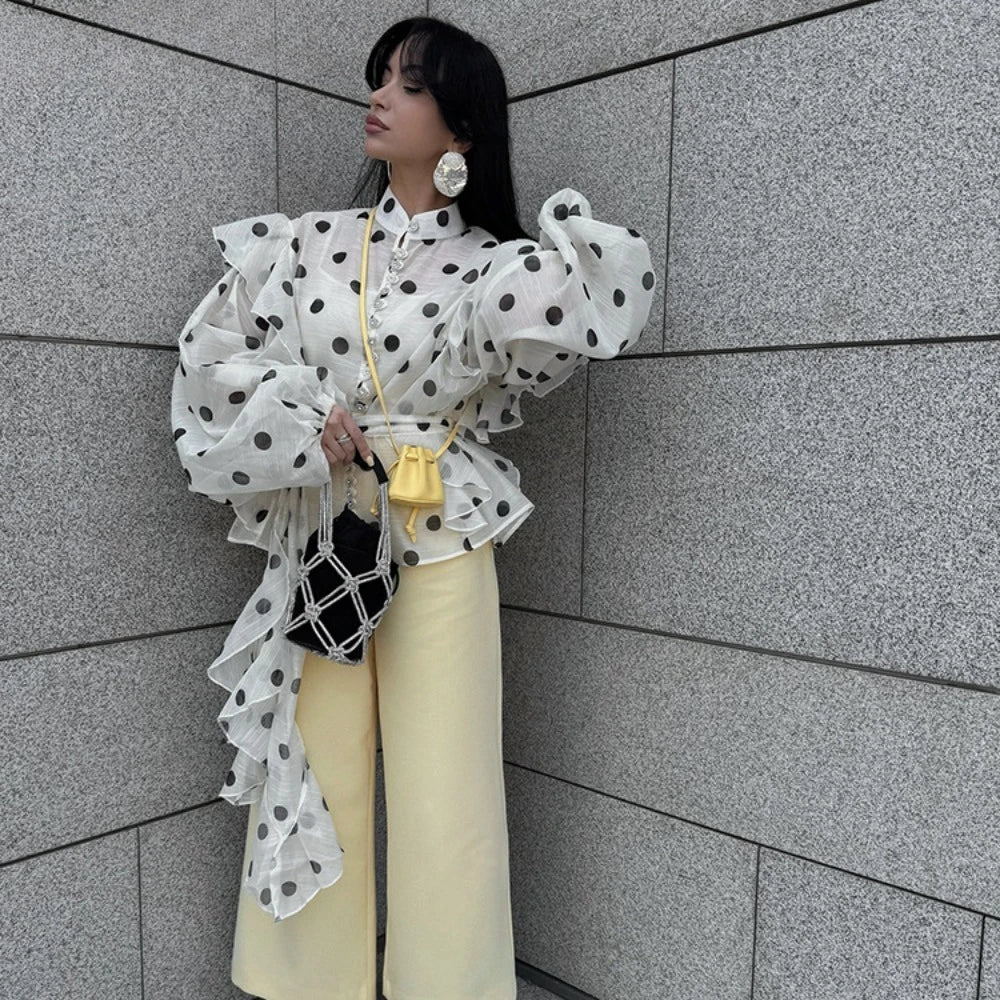 Person wearing a polka dot blouse and yellow pants against a gray tiled wall.